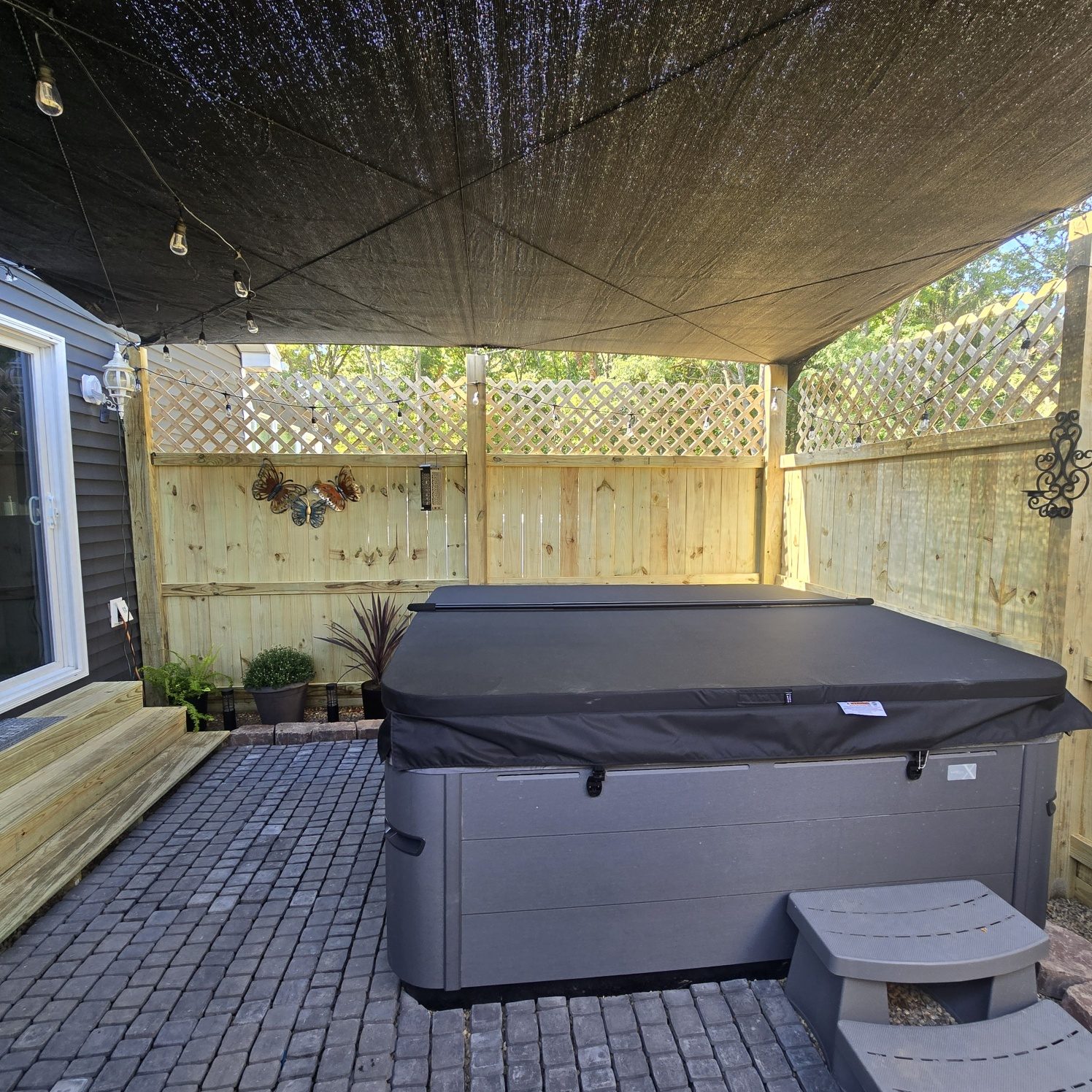 Finished backyard hot tub retreat with paver patio, privacy fence, lattice, shade sail and string lights in Southwest Michigan